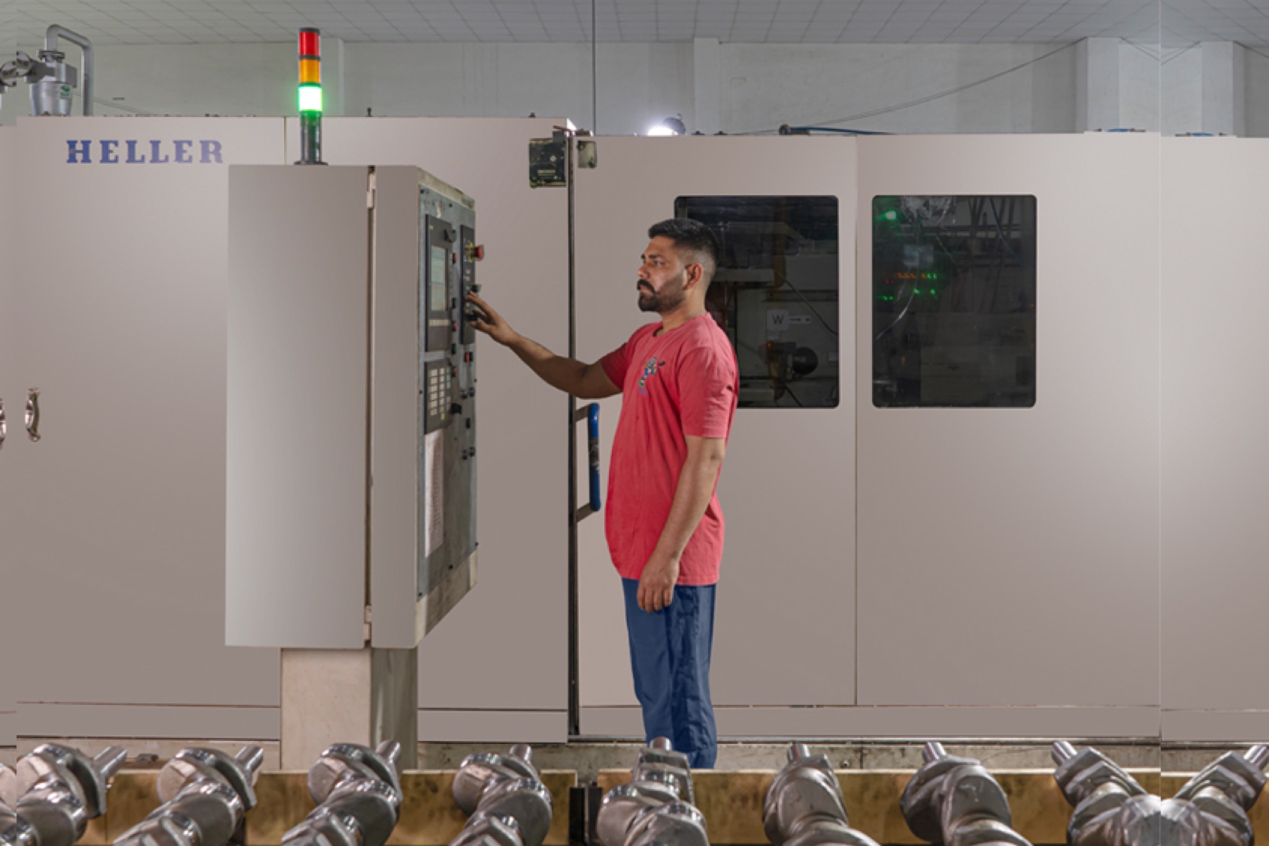 Machine operator at the control panel of a Heller machining centre used for crank pin milling, with finished crankshafts racked in front